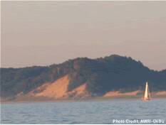 A forested sand dune at sunset sits adjacent to a lake, with a sailboat passing by.